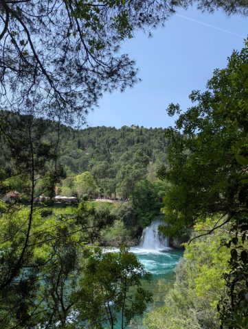 cascada-krka-vedere-prin-frunzis-parc-national-croatia Vedere prin frunziș către o cascadă din Parcul Național Krka, Croația, cu dealuri verzi în fundal.