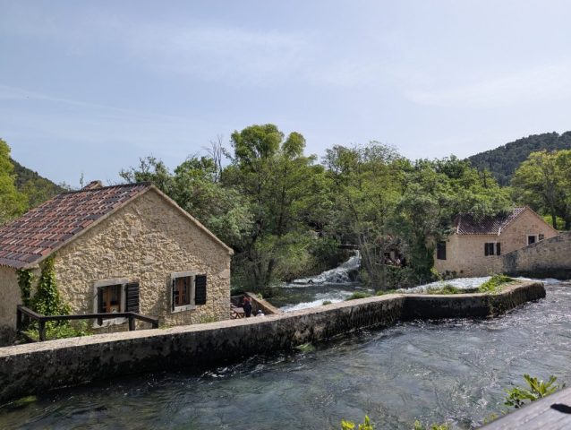 case-traditionale-krka-croatia-natura-apa Case tradiționale din piatră în Parcul Național Krka, Croația, lângă apa curgătoare și pădure.